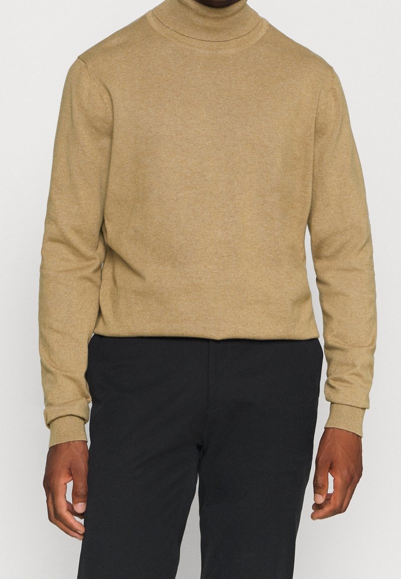 Homme portant un pull à col roulé beige et un pantalon noir, debout les mains détendues le long du corps devant un fond uni.