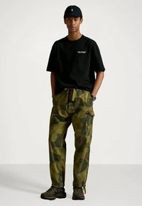 Ung man som bär en svart Polo Sport T-shirt, svart keps, kamouflage cargobyxor och svarta sneakers, står med händerna i fickorna.