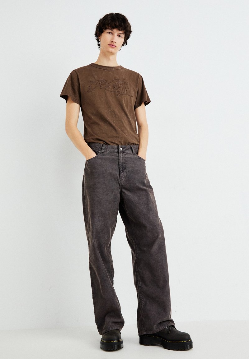 T-shirt in cotone marrone con logo in rilievo, abbinato a pantaloni larghi grigi scuri. Stivaletti neri completano l'outfit.