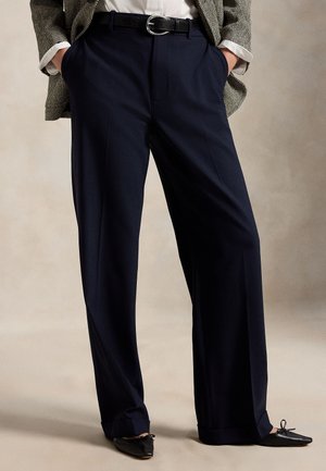 Persona che indossa pantaloni larghi blu navy con cintura nera, una camicetta bianca, un blazer a spina di pesce e ballerine nere a punta. Mani nelle tasche.