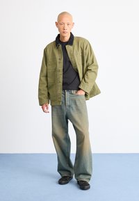 Olive green utility jacket with black collar, worn over a black shirt, paired with wide light blue jeans and black shoes on a blue floor.