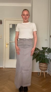 Gonna grigia lunga con vita alta e una texture sottile, abbinata a una t-shirt bianca aderente. Stivali neri completano l'outfit.