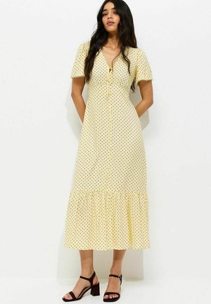 Kobieta ubrana w żółtą sukienkę midi w czarne groszki, z krótkimi rękawami, z przodu z guzikami i falbaną na dole, w połączeniu z czarnymi sandałami na słupku.