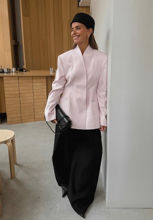 Sieviete, kas smaida, tērpusies gaismas rozā strukturētā žaketē, melnās plašās biksēs, melnā cepurē un tur rokās melnu maku, stāv telpā pie baltas sienas.