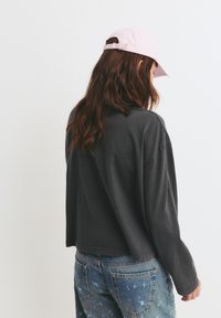 Camicia lunga grigia over-size con orlo cropped, abbinata a jeans in denim blu strappati e un cappellino da baseball rosa chiaro.