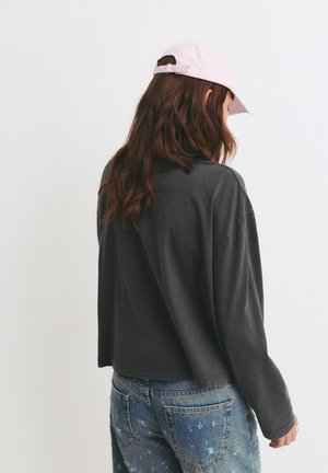 Grijs oversized langemouwen shirt met een cropped zoom, gecombineerd met beschadigde blauwe spijkerbroek en een lichtroze baseballcap.