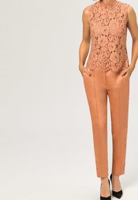 Pfirsichfarbener Spitzen-Ärmelloser Oberteil mit gewelltem Saum, kombiniert mit maßgeschneiderten pfirsichfarbenen Hosen und schwarzen spitzen High Heels. Glatte Textur.
