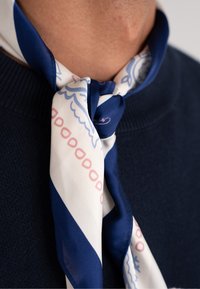 Foulard di seta con una base bianca e strisce diagonali blu e accenti rosa, annodato intorno al collo, poggiato su un maglione blu navy.
