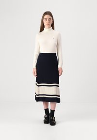 Pull à col roulé blanc et jupe midi plissée bleu marine avec des rayures crème et noires. Sandales noires avec boucles. Design simple et ajusté.