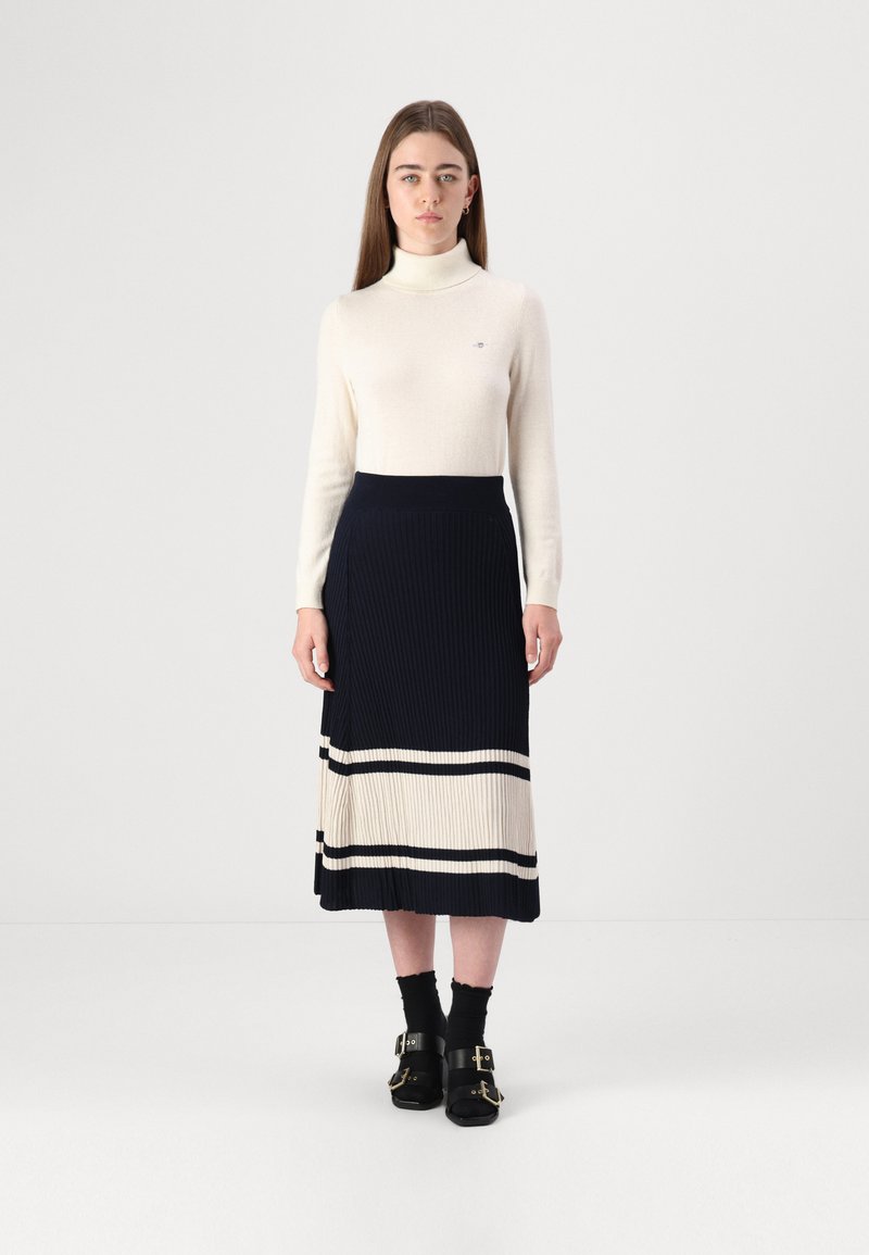 Pull à col roulé blanc et jupe midi plissée bleu marine avec des rayures crème et noires. Sandales noires avec boucles. Design simple et ajusté.