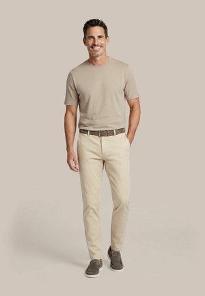 Man staat, glimlacht, draagt een beige T-shirt, lichtbeige broek met een bruine riem en bruine instapschoenen, tegen een beige achtergrond.