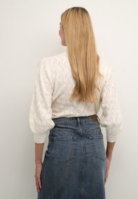 Pull en tricot couleur crème avec des manches bouffantes, présentant un motif texturé. Associé à une jupe en denim sombre avec des coutures contrastantes.