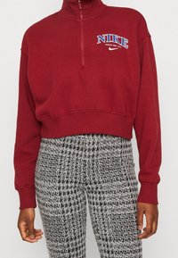 Burgundy Nike-tröja med hög krage och kvartsgångdragkedja. Har en vit och blå logotyp på bröstet. Kombinerad med texturerade svarta och vita mönstrade byxor.