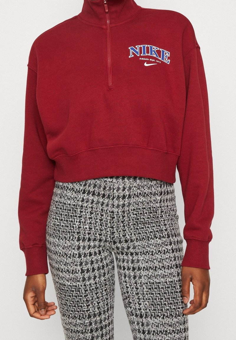 Burgundy Nike-tröja med hög krage och kvartsgångdragkedja. Har en vit och blå logotyp på bröstet. Kombinerad med texturerade svarta och vita mönstrade byxor.