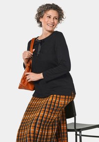 Schwarzes Langarmshirt mit einer Schleifen-Detail, kombiniert mit einem gemusterten orange- und schwarzkarierter Rock. Das Modell hält eine braune Lederhandtasche.
