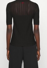 Top in maglia nera a coste con maniche corte, presenta un pannello trasparente sul retro e un orlo ondulato. Include un piccolo etichetta rossa come accento sul collo.
