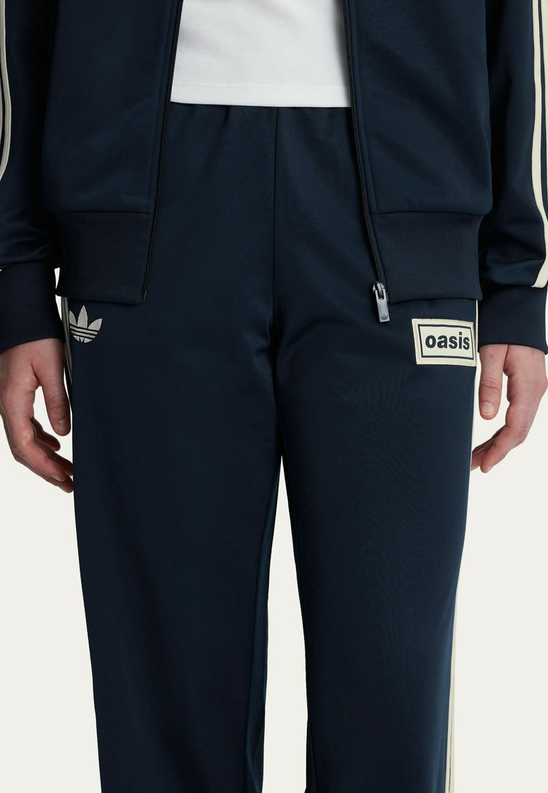 Navy tracksuit bottom with white accent stripes, featuring an "oasis" patch and Adidas logo in light gray on the left thigh. Zippered pocket included.