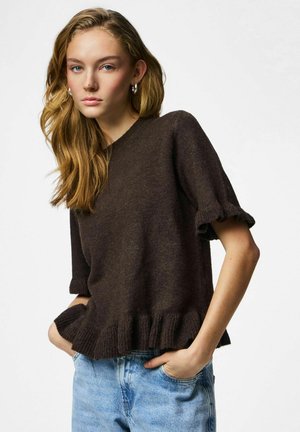 Jeune femme aux longs cheveux blonds portant un pull à manches courtes marron foncé et un jean bleu clair, debout les mains dans les poches.