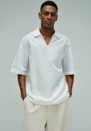 Jeune homme aux cheveux courts portant une chemise blanche à manches courtes avec col et un pantalon beige, debout les mains dans les poches devant un fond uni.