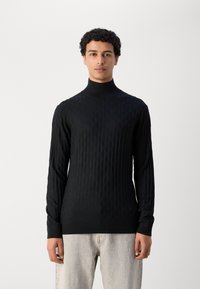 Schwarzer Rollkragenpullover mit strukturiertem Rautenmuster, langen Ärmeln und figurbetontem Design. Kombiniert mit hellgrauen Jeans.