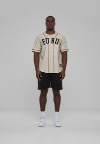 Maglietta da baseball beige con "FUBU" in lettere nere, accentuata da dettagli neri; abbinata a pantaloni corti neri e sneakers bianche.