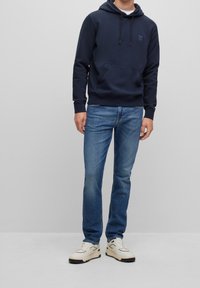 Navy hooded sweatshirt with front pocket, cotton blend material. Paired with light blue jeans and white sneakers with black accents.