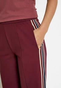 Bordeauxkleurige joggingsbroek met een ontspannen pasvorm, voorzien van zijzakken en verticale strepen in crème en marineblauw langs de naden. Zachte textuur.