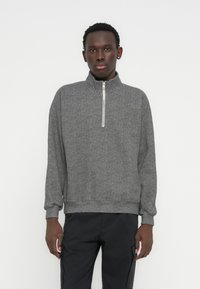 Sweat-shirt gris à fermeture éclair avec un col haut, présentant un motif chevrons texturé, des poignets et un ourlet côtelés, associé à un pantalon foncé.