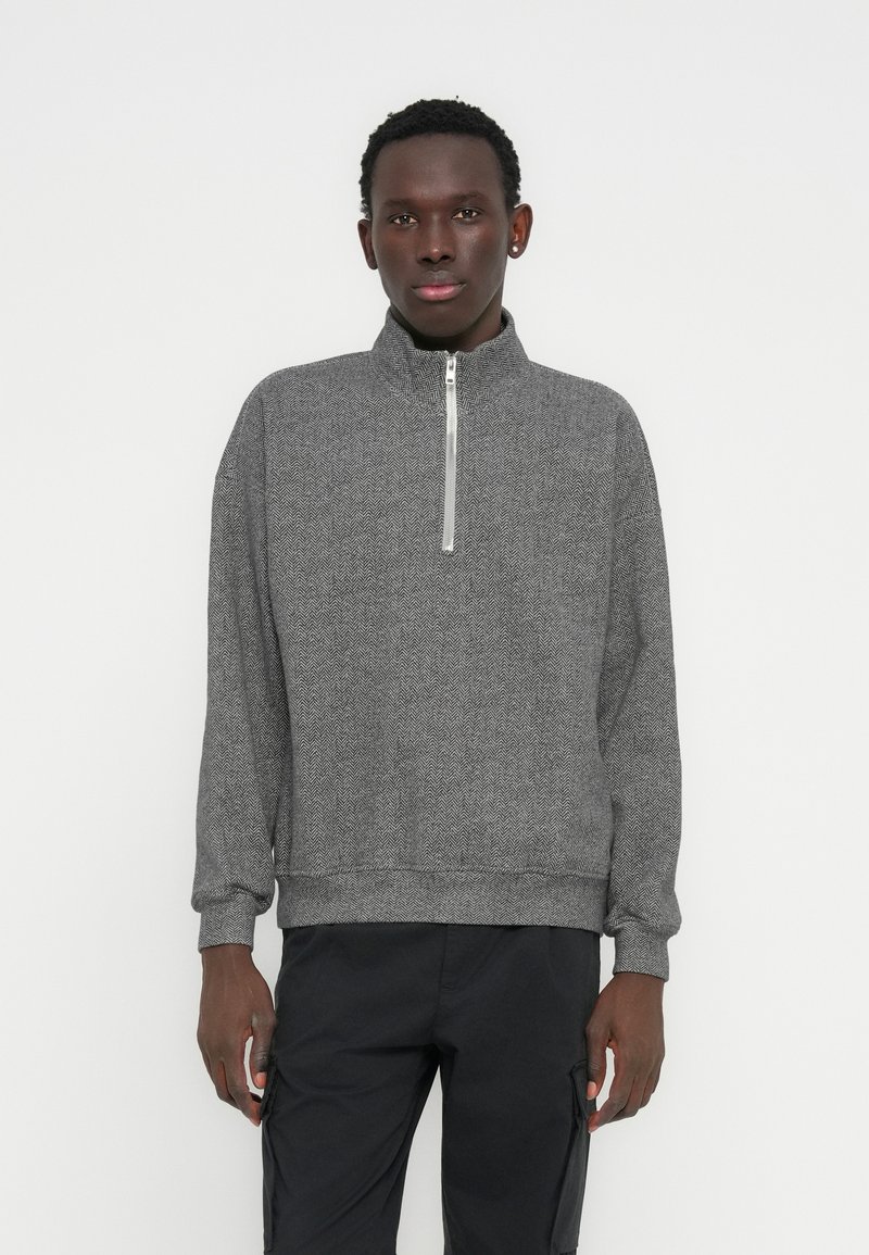 Sweat-shirt gris à fermeture éclair avec un col haut, présentant un motif chevrons texturé, des poignets et un ourlet côtelés, associé à un pantalon foncé.