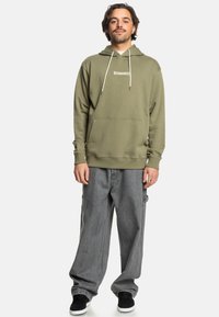 Olijfgroen hoodie met een voorzak, trekkoorden en een wit logo; gecombineerd met losse grijze jeans en zwarte schoenen. Zachte stofstructuur.