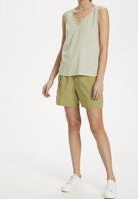 Top senza maniche verde chiaro con rifiniture in pizzo, abbinato a pantaloni corti verde oliva chiaro. La modella indossa sneakers bianche, in piedi su uno sfondo neutro.