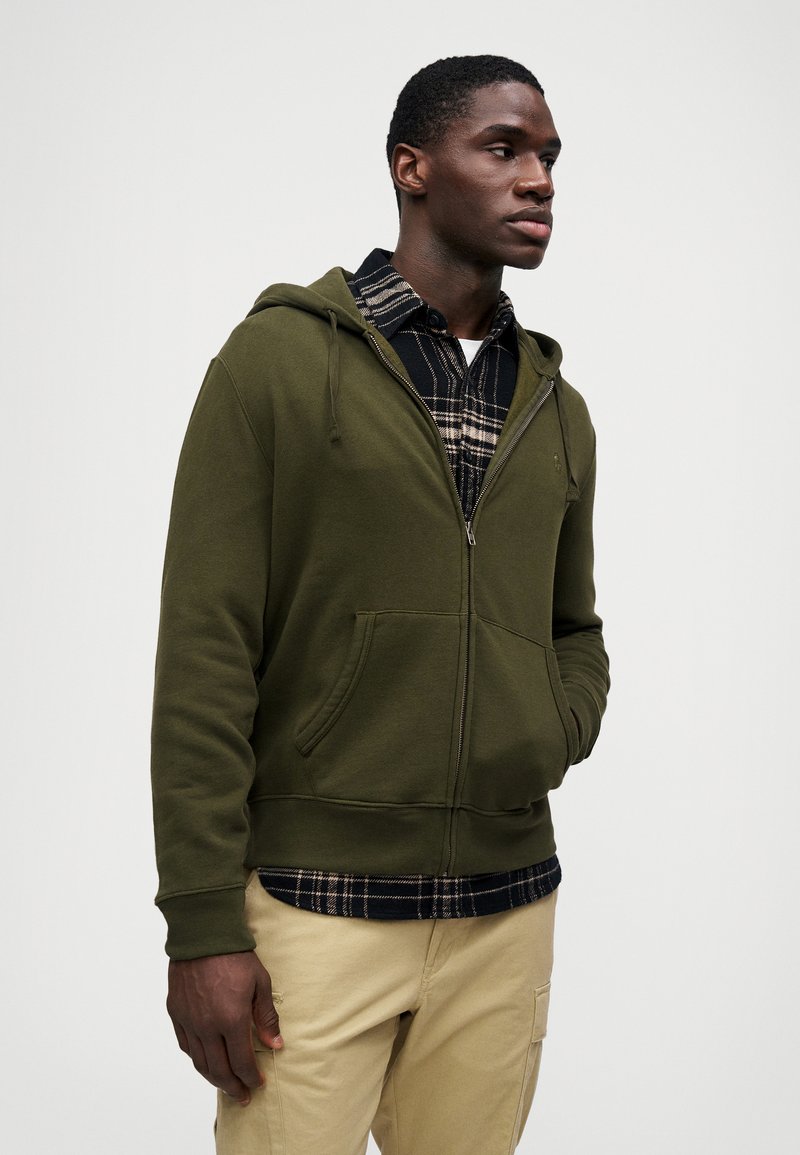 Sudadera con cremallera en verde oliva, con un bolsillo frontal y capucha ajustable, llevada sobre una camisa de cuadros en blanco y negro, combinada con pantalones beige.