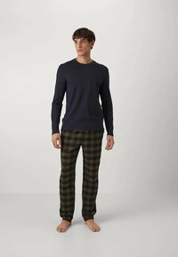 Άνδρας που στέκεται γυμνοπόδος φορώντας μπλε μακρυμάνικη μπλούζα και σκούρο πράσινο-μαύρο καρό παντελόνι, μπροστά σε ένα απλό φωτεινό φόντο.