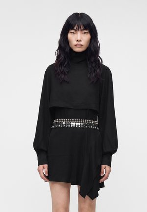 Femme aux longs cheveux noirs portant une robe noire à manches longues avec un col montant et une ceinture cloutée brillante autour de la taille.