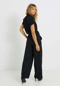 Combinaison noire à manches courtes, taille à cordon de serrage, design à jambe large et tissu léger. Portée avec des chaussures à talons hauts, faisant face à l'arrière.