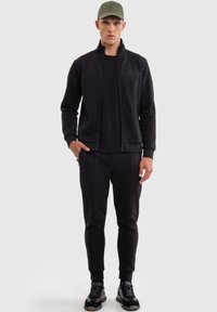 Chaqueta negra con cremallera y pantalones a juego en tejido suave, con un silueta ajustada, puños acanalados y bolsillos laterales, combinados con zapatillas negras.