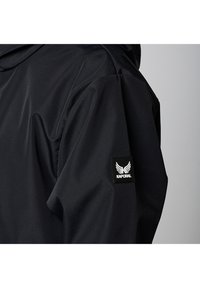 Chaqueta negra con una textura lisa y mate. Presenta un parche del logo KAPORAL en el hombro izquierdo y una capucha. Diseño ajustado con una forma minimalista.