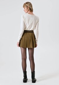 Maglia a maniche lunghe color crema, gonna plissettata verde oliva con una cintura scura, stivali neri alla caviglia e collant neri trasparenti. Texture liscia, design su misura.