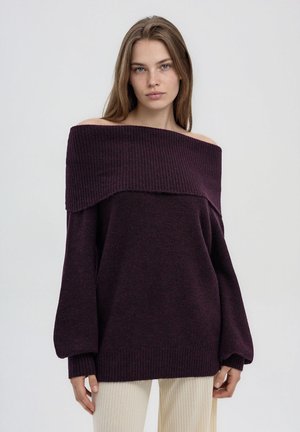 Giovane donna con lunghi capelli castani che indossa un maglione oversize viola scuro a spalle scoperte e pantaloni beige chiaro su uno sfondo neutro.