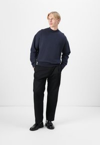 Tengerész kereknyakú pulóver fekete nadrággal párosítva. Sima szövettextúra, laza fazon és minimális dizájn. Lábbeli fekete bőrcipő.