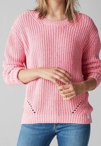 Jasnoróżowy sweter dzianinowy z prążkowaną teksturą, okrągłym dekoltem i bocznymi rozcięciami zapewniającymi wentylację. Wykończony małą metką na dolnym brzegu.
