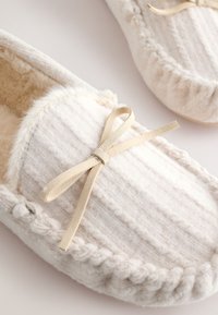 Pa paire de chaussons tricotés couleur crème doublés en fausse fourrure avec des accents de nœuds en daim beige sur un fond clair.