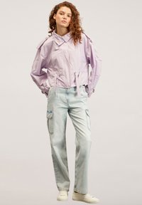 Giacca corta viola chiaro con vita regolabile, vestibilità oversize e dettagli con bottoni; abbinata a jeans cargo blu chiaro e sneakers bianche.
