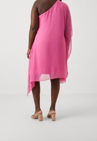 Femme portant une robe rose à une épaule et des sandales à talons blocs ouvertes beiges, se tenant contre un fond uni clair.