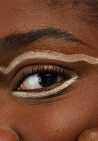 Gros plan sur un œil à la peau foncée avec un eye-liner blanc audacieux créant une forme ondulée. Les cils sont longs, avec une texture de peau lisse entourant l'œil.