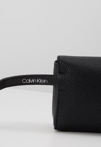 Černá kožená taštička s texturovaným povrchem, zaoblenými okraji a popruhem se bílým logem „Calvin Klein“. Jednoduchý, kompaktní design.