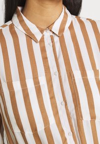 Primo piano di una camicia button-up a strisce verticali marroni e bianche con due tasche frontali e colletto indossata da una persona.