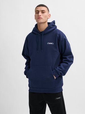 Jonge man met een marineblauwe hummel hoodie en zwarte hummel broek, staand met handen in de capuchonzak tegen een effen achtergrond.