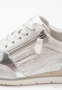 Srebrno-białe buty sportowe wykonane z mieszanych materiałów, z fakturowaną cholewką z zamszu, zapięciem na sznurowadła, bocznym zamkiem oraz lekką gumową podeszwą.