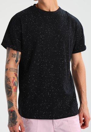 Czarny bawełniany T-shirt z nakrapianym białym wzorem. Okrągły dekolt, krótkie rękawy, luźny krój. Charakteryzuje się widocznymi tatuażami na przedramionach.